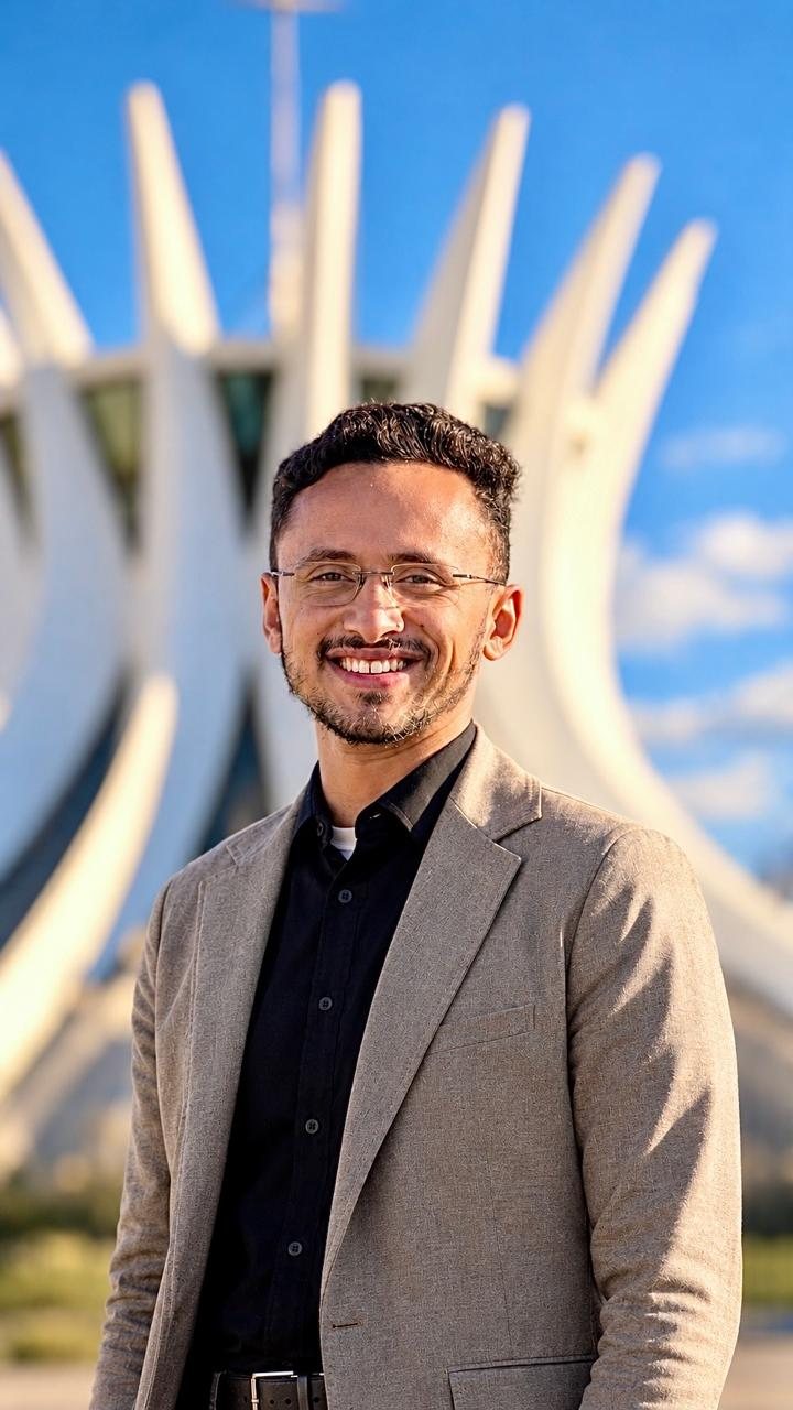 Vinicius Nascimento sorrindo, de blazer bege, em frente a uma catedral de arquitetura moderna
